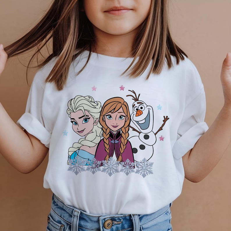 May include: White t-shirt featuring Elsa, Anna, and Olaf from the movie Frozen. The graphic includes the characters' faces, snowflakes, and stars. The shirt is worn by a person.