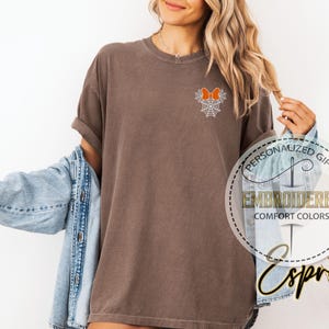 Pu&ograve; includere: T-shirt marrone con un ricamo di un fiocco arancione e una ragnatela. La maglietta &egrave; del marchio Comfort Colors. Il testo sulla maglietta dice "Embroidered Comfort Colors".