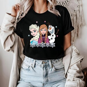 May include: Black t-shirt featuring a graphic of Elsa, Anna, and Olaf from Frozen. The design includes snowflakes and stars. The characters are depicted in their signature outfits, with Elsa in a blue dress and Anna in a purple cape.