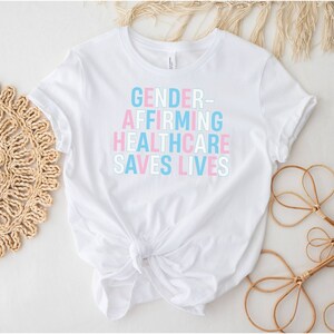 May include: White t-shirt with the text "GENDER-AFFIRMING HEALTHCARE SAVES LIVES" in pink and blue lettering.