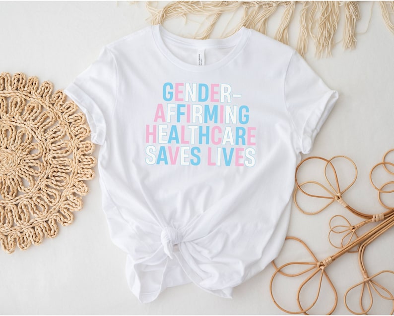 May include: White t-shirt with the text "GENDER-AFFIRMING HEALTHCARE SAVES LIVES" in pink and blue lettering.