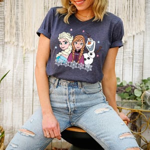 May include: Navy blue t-shirt featuring a graphic of Elsa, Anna, and Olaf from the movie Frozen. The design includes snowflakes and pink stars. The shirt has rolled-up sleeves and is paired with light blue ripped jeans.