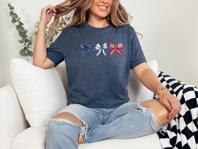 May include: A navy blue t-shirt featuring three embroidered bows in patriotic colors: blue with white polka dots, white, and red with white stripes. The shirt is worn with ripped blue jeans.