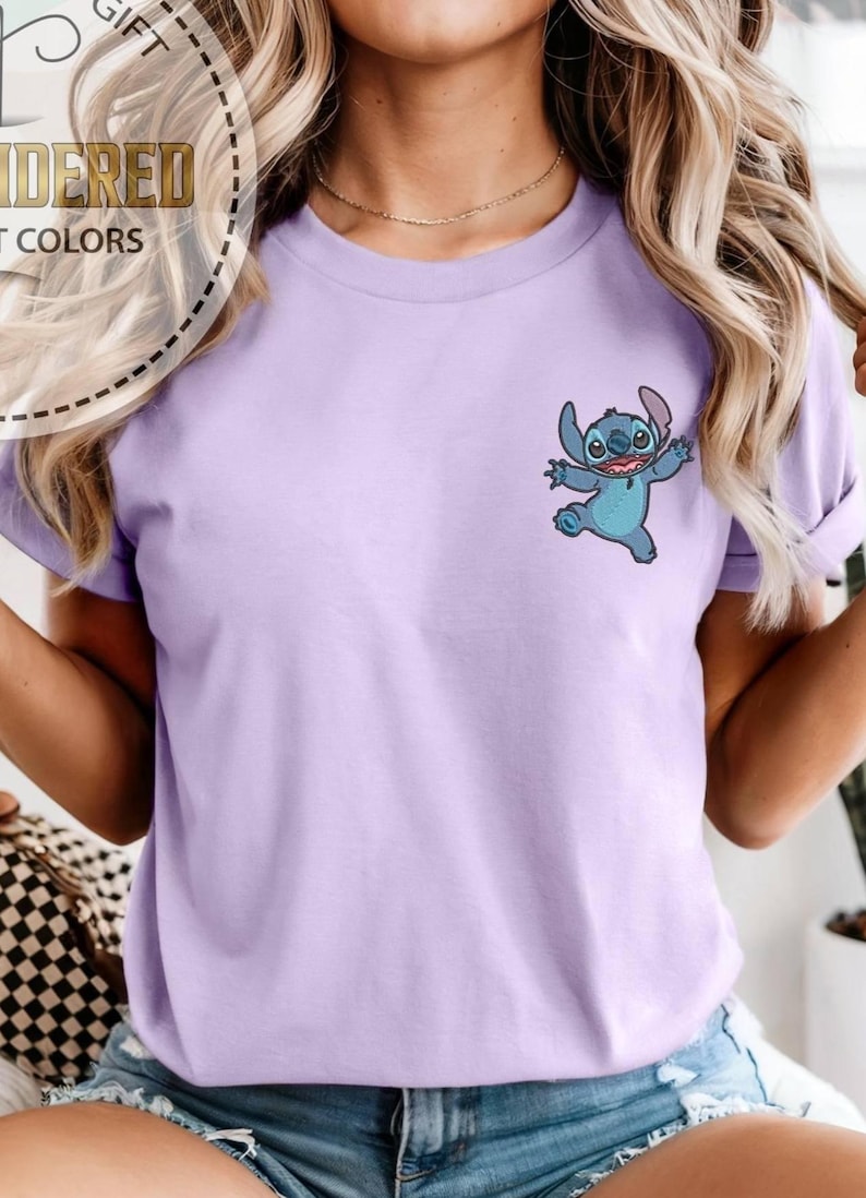 May include: A light purple t-shirt featuring a small embroidered image of the character Stitch from the animated film Lilo & Stitch. The cartoon character is blue with large eyes and is in a playful pose. The shirt has short sleeves.