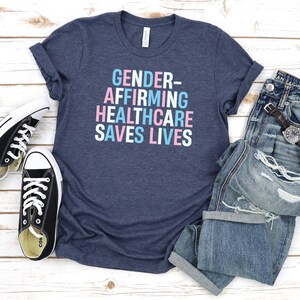 May include: A heather navy blue t-shirt with the text "GENDER-AFFIRMING HEALTHCARE SAVES LIVES" in pink and blue lettering.