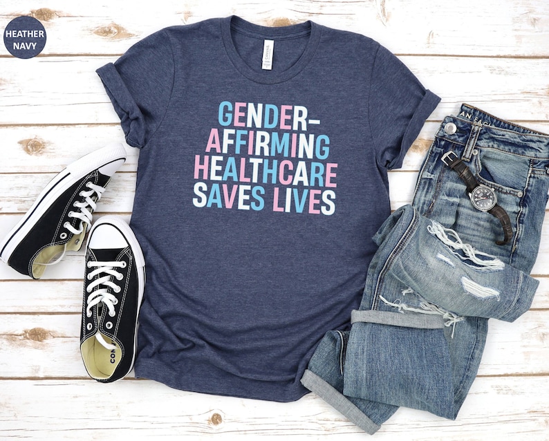 May include: A heather navy blue t-shirt with the text "GENDER-AFFIRMING HEALTHCARE SAVES LIVES" in pink and blue lettering.