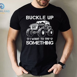 Op de afbeelding: Zwart T-shirt met een witte afbeelding van een jeep met de tekst "Buckle Up I Want To Try Something"