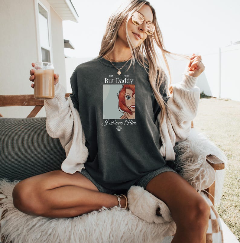 Może przedstawiać: Ciemnoszara koszulka unisex z grafiką rudowłosej postaci z kresk&oacute;wki i napisem "But Daddy I Love Him". Koszulka ma luźny kr&oacute;j i wygląd vintage.