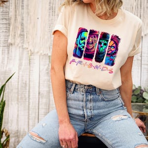 May include: Cream-colored t-shirt featuring a graphic of four horror movie characters in vibrant colors. The word "FRIENDS" is written in pink and purple below the characters. The person is wearing blue jeans.