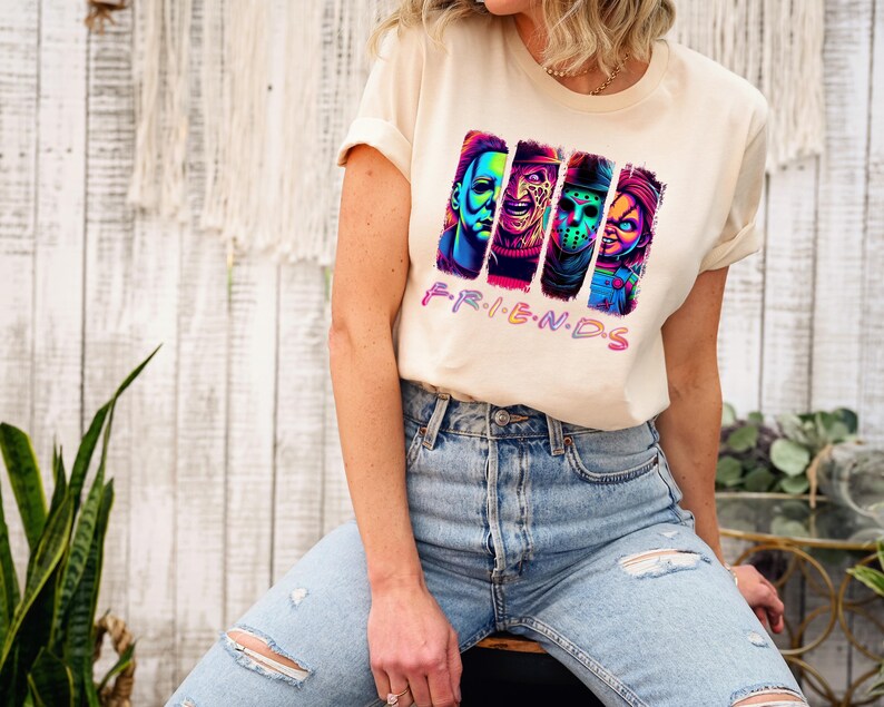 May include: Cream-colored t-shirt featuring a graphic of four horror movie characters in vibrant colors. The word "FRIENDS" is written in pink and purple below the characters. The person is wearing blue jeans.