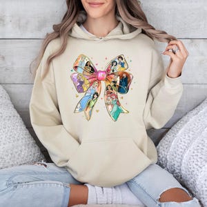 May include: Beige hoodie featuring a large bow design with various princess illustrations. The bow is filled with colorful images of Disney princesses. The hoodie has a front pocket and a drawstring hood.