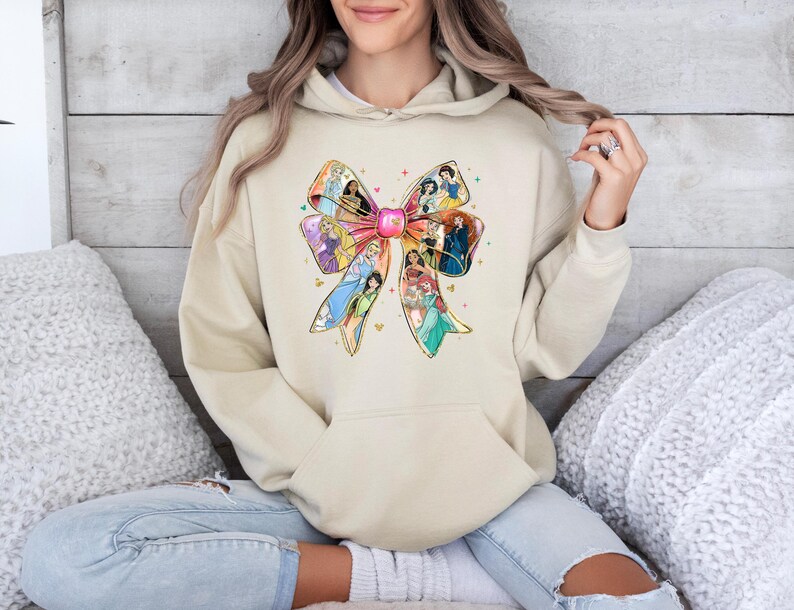 May include: Beige hoodie featuring a large bow design with various princess illustrations. The bow is filled with colorful images of Disney princesses. The hoodie has a front pocket and a drawstring hood.