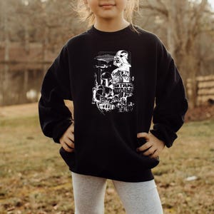 May include: Black sweatshirt featuring a white graphic of Star Wars characters and the Star Wars logo. The design includes Darth Vader, a Stormtrooper, and other iconic elements from the film series. The sweatshirt is worn by a person.