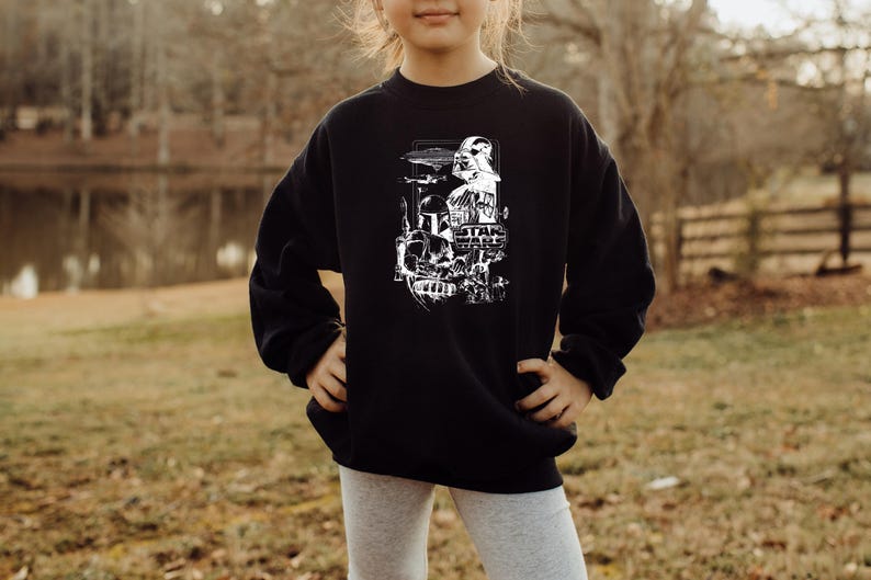 May include: Black sweatshirt featuring a white graphic of Star Wars characters and the Star Wars logo. The design includes Darth Vader, a Stormtrooper, and other iconic elements from the film series. The sweatshirt is worn by a person.