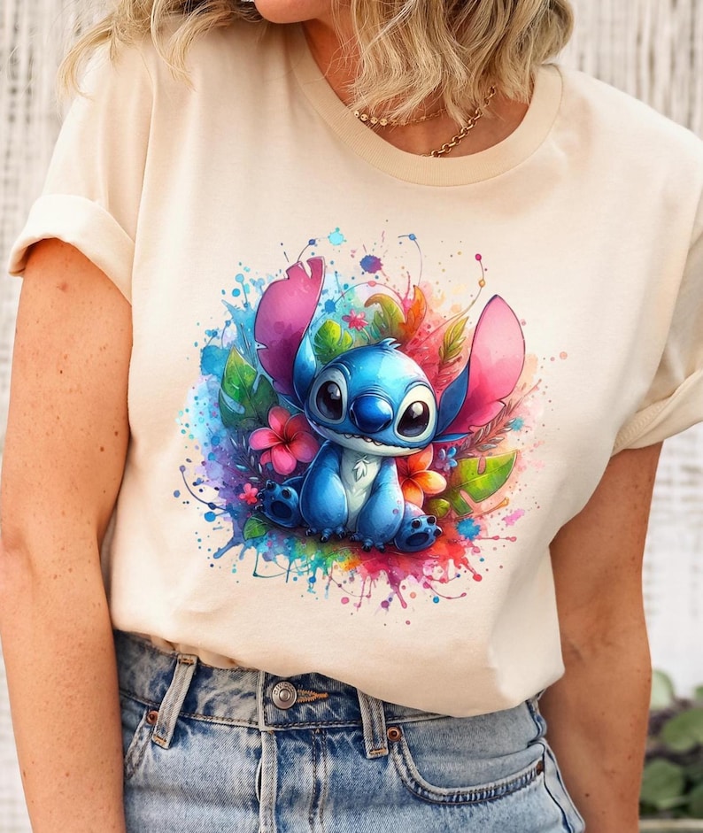 Op de afbeelding: Cr&egrave;mekleurig T-shirt met een aquarel illustratie van een blauw cartoonfiguur met grote oren, omringd door kleurrijke bloemen en spetters. Het personage is het middelpunt, tegen een levendige, artistieke achtergrond.
