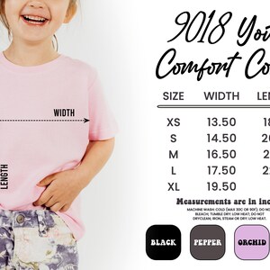 May include: A light pink youth t-shirt with size and measurement details. The shirt's width ranges from 13.50 to 19.50 inches, and the length from 18.75 to 26 inches. Available colors include black, pepper, orchid, and blossom.