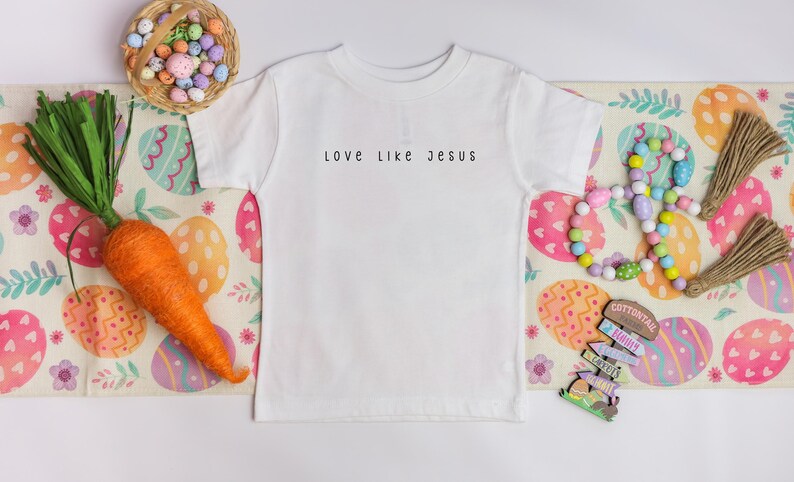 May include: A white t-shirt with the text "love like Jesus" in black. The shirt is on a decorative Easter-themed runner with a carrot, basket of eggs, and wooden beads.