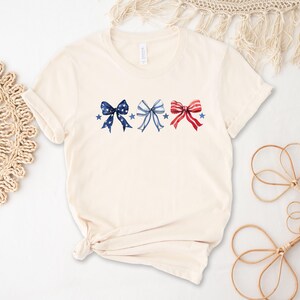 May include: Cream-colored t-shirt featuring three watercolor bows in patriotic colors: navy blue, light blue, and red. Each bow is adorned with stars. The shirt has short sleeves and a crew neck.