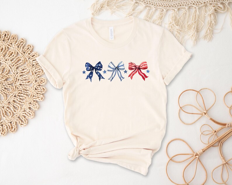 May include: Cream-colored t-shirt featuring three watercolor bows in patriotic colors: navy blue, light blue, and red. Each bow is adorned with stars. The shirt has short sleeves and a crew neck.