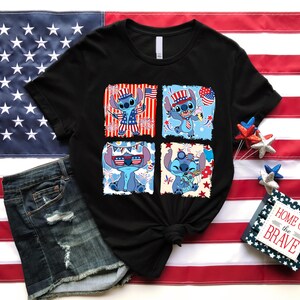 May include: Black t-shirt featuring four panels with a cartoon character in patriotic attire. The character is blue with large ears and is wearing sunglasses, a hat, and holding a flag. The shirt is displayed with denim shorts and an American flag.
