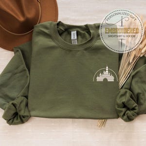 May include: Olive green sweatshirt with a white castle graphic. The sweatshirt has long sleeves and a crew neck. A brown hat and decorative wheat stalks are also in the image. The text "PERSONALIZED GIFT EMBROIDERED SWEATSHIRT & HOODIE" is in a circle.