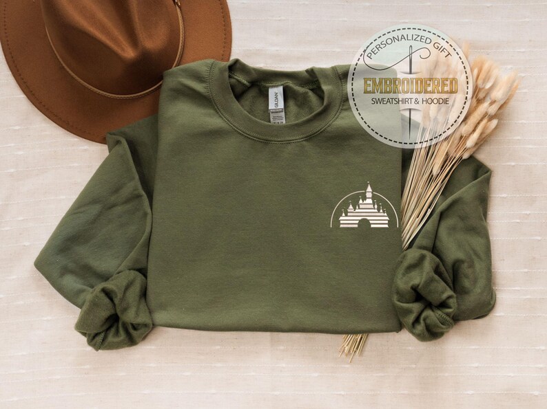 May include: Olive green sweatshirt with a white castle graphic. The sweatshirt has long sleeves and a crew neck. A brown hat and decorative wheat stalks are also in the image. The text "PERSONALIZED GIFT EMBROIDERED SWEATSHIRT & HOODIE" is in a circle.