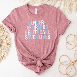 May include: A heather mauve t-shirt with the text "GENDER-AFFIRMING HEALTHCARE SAVES LIVES" printed in blue, pink, and white.