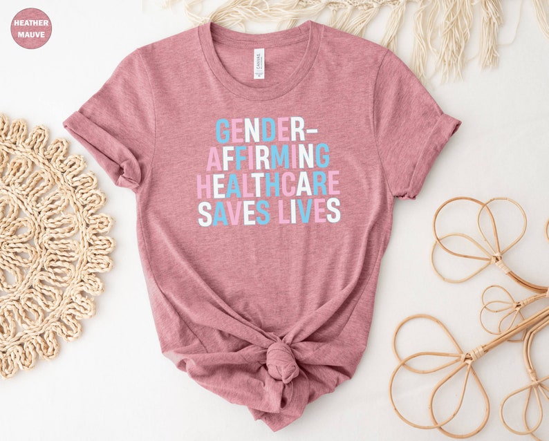 May include: A heather mauve t-shirt with the text "GENDER-AFFIRMING HEALTHCARE SAVES LIVES" printed in blue, pink, and white.