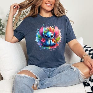 Op de afbeelding: Marineblauw t-shirt met een kleurrijke afbeelding van een blauw stripfiguur omringd door bloemen en verfspatten. Het ontwerp is gecentreerd op het shirt, wat zorgt voor een levendige en speelse uitstraling. Het shirt is gemaakt van zacht materiaal.