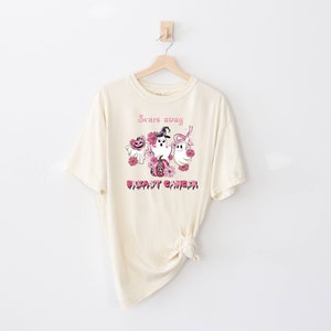 Pode incluir: Uma camiseta bege com uma fita rosa e o texto "Scare away breast cancer" com um gr&aacute;fico de fantasmas e ab&oacute;boras.