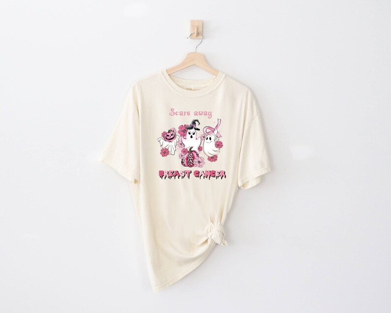 Pode incluir: Uma camiseta bege com uma fita rosa e o texto "Scare away breast cancer" com um gr&aacute;fico de fantasmas e ab&oacute;boras.