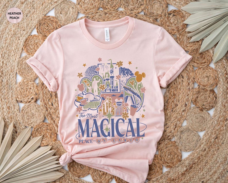 May include: Heather peach-colored t-shirt with a crew neck and rolled sleeves. The front features a colorful graphic of a castle, fireworks, and other park icons, with the text "The Most Magical Place on Earth."