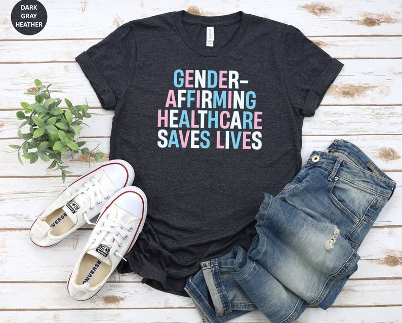 May include: Dark heather gray t-shirt with the text "GENDER-AFFIRMING HEALTHCARE SAVES LIVES" in white letters with pink and blue accents.