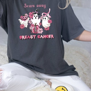 Pode incluir: Uma camiseta cinza escura com o texto "Scare away breast cancer" em letras cor-de-rosa que pingam. O design apresenta tr&ecirc;s fantasmas com flores cor-de-rosa e uma fita cor-de-rosa. Um fantasma est&aacute; usando um chap&eacute;u de bruxa.