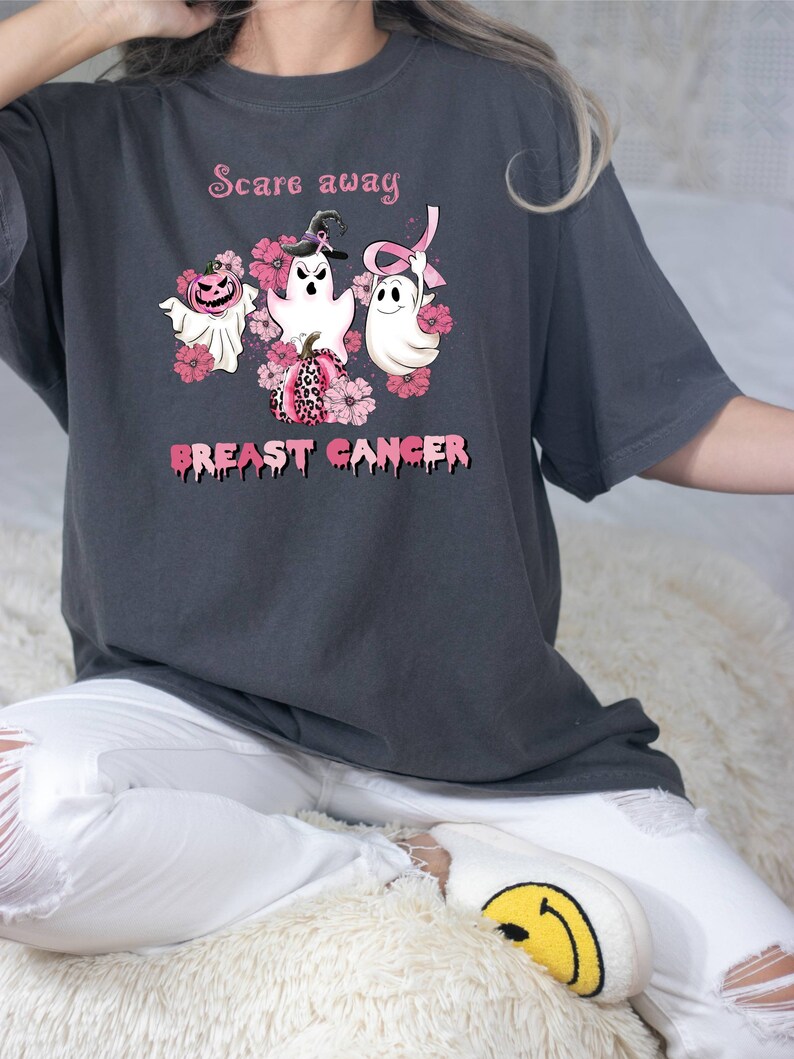 Pode incluir: Uma camiseta cinza escura com o texto "Scare away breast cancer" em letras cor-de-rosa que pingam. O design apresenta tr&ecirc;s fantasmas com flores cor-de-rosa e uma fita cor-de-rosa. Um fantasma est&aacute; usando um chap&eacute;u de bruxa.