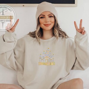 Puede incluir: Sudadera beige con un dise&ntilde;o de castillo bordado y las palabras "Disney World" en amarillo. La sudadera tiene mangas largas y cuello redondo. Se lleva un gorro beige en la cabeza.