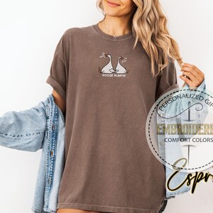 Pu&ograve; includere: T-shirt marrone con disegno di oca ricamato e la scritta "GOOSE BUMPS!". La maglietta &egrave; di un colore confortevole e ha le maniche arrotolate. Una giacca di jeans &egrave; drappeggiata sulle spalle.