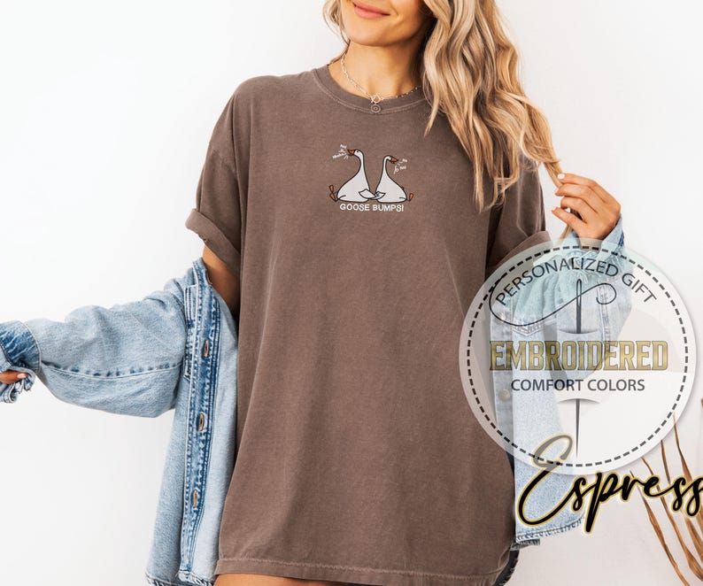 Pu&ograve; includere: T-shirt marrone con disegno di oca ricamato e la scritta "GOOSE BUMPS!". La maglietta &egrave; di un colore confortevole e ha le maniche arrotolate. Una giacca di jeans &egrave; drappeggiata sulle spalle.