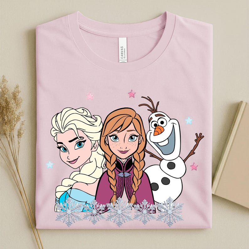 May include: A light pink t-shirt featuring Elsa, Anna, and Olaf from the movie Frozen. The graphic includes the characters with snowflakes and stars. The shirt is folded and has a "CANVAS" tag.