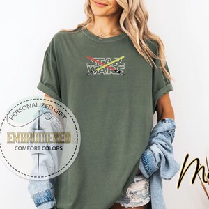 Może przedstawiać: Zielona koszulka z haftem "STAR WARS" z przodu. Tekst jest biały z czerwonymi i ż&oacute;łtymi ostrzami miecza świetlnego krzyżującymi się z tyłu.