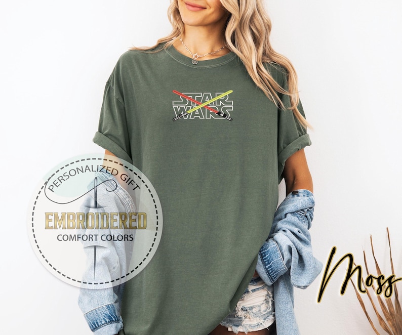 Może przedstawiać: Zielona koszulka z haftem "STAR WARS" z przodu. Tekst jest biały z czerwonymi i ż&oacute;łtymi ostrzami miecza świetlnego krzyżującymi się z tyłu.