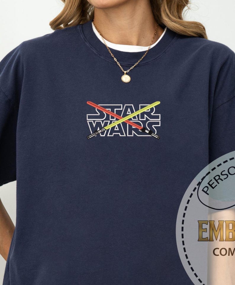 Może przedstawiać: Koszulka w kolorze granatowym z białym obrysem sł&oacute;w "STAR WARS" i dwoma skrzyżowanymi mieczami świetlnymi w kolorze czerwonym i ż&oacute;łtym.