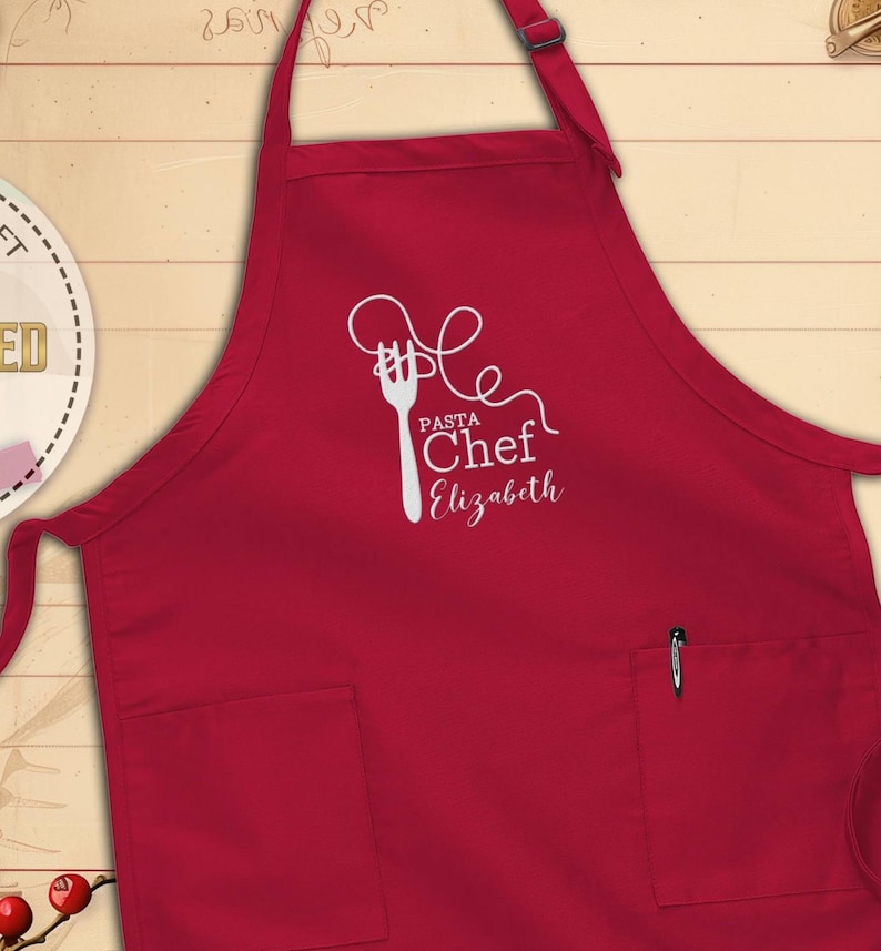 May include: A red apron with a white graphic of a fork and pasta, with the words "Pasta Chef Elizabeth" in a script font. The apron has two front pockets and a pen in the right pocket.