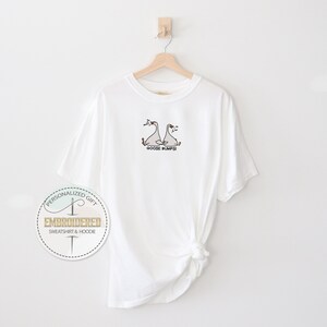 Pu&ograve; includere: T-shirt bianca con un ricamo di due oche e la scritta "Goose Bumps!" in nero. La maglietta &egrave; appesa a una gruccia di legno contro un muro bianco. Un logo circolare &egrave; visibile in basso a sinistra.