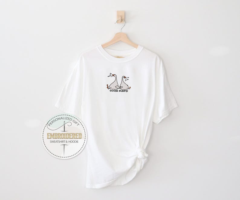 Pu&ograve; includere: T-shirt bianca con un ricamo di due oche e la scritta "Goose Bumps!" in nero. La maglietta &egrave; appesa a una gruccia di legno contro un muro bianco. Un logo circolare &egrave; visibile in basso a sinistra.