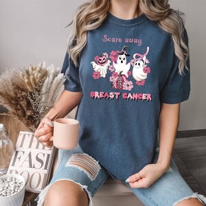 Pode incluir: Uma camiseta azul marinho com uma fita rosa, tr&ecirc;s fantasmas e ab&oacute;boras. O texto na camiseta diz "Scare away breast cancer".