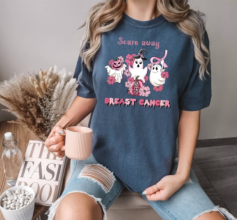 Pode incluir: Uma camiseta azul marinho com uma fita rosa, tr&ecirc;s fantasmas e ab&oacute;boras. O texto na camiseta diz "Scare away breast cancer".