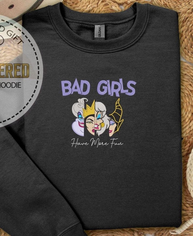 Puede incluir: Sudadera negra con el texto "BAD GIRLS" en morado sobre un gr&aacute;fico de tres caras de villanas de dibujos animados. Debajo, la frase "Have More Fun" en cursiva. La sudadera es de la marca Gildan.