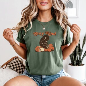 May include: Green t-shirt with the text "Stay Kind" in orange script. The shirt features a graphic of a dark brown monkey sitting on a brown teddy bear. The shirt has short sleeves.