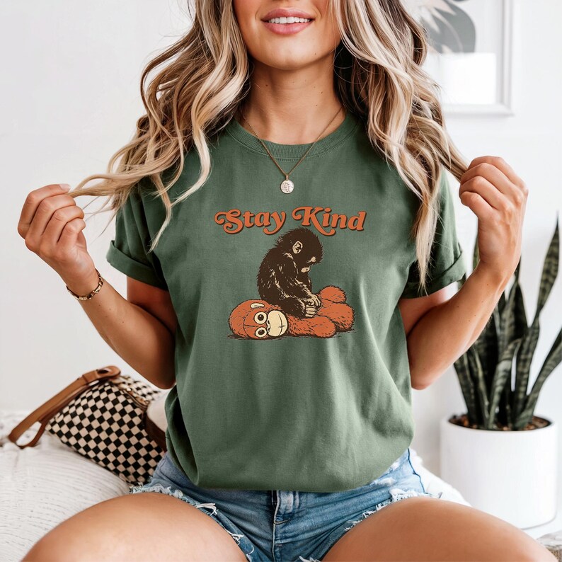 May include: Green t-shirt with the text "Stay Kind" in orange script. The shirt features a graphic of a dark brown monkey sitting on a brown teddy bear. The shirt has short sleeves.